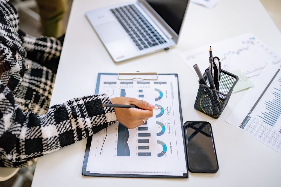 An office setting with a person analyzing charts and graphs at a desk with a laptop and smartphone.